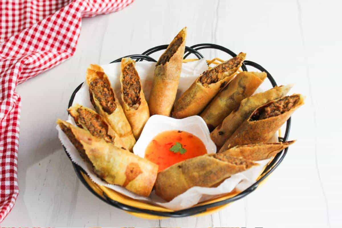 A black basket filled with cut spring rolls is arranged in a circle on a white surface. A small square dish in the center contains orange dipping sauce. A red and white checkered cloth is partially visible on the left.