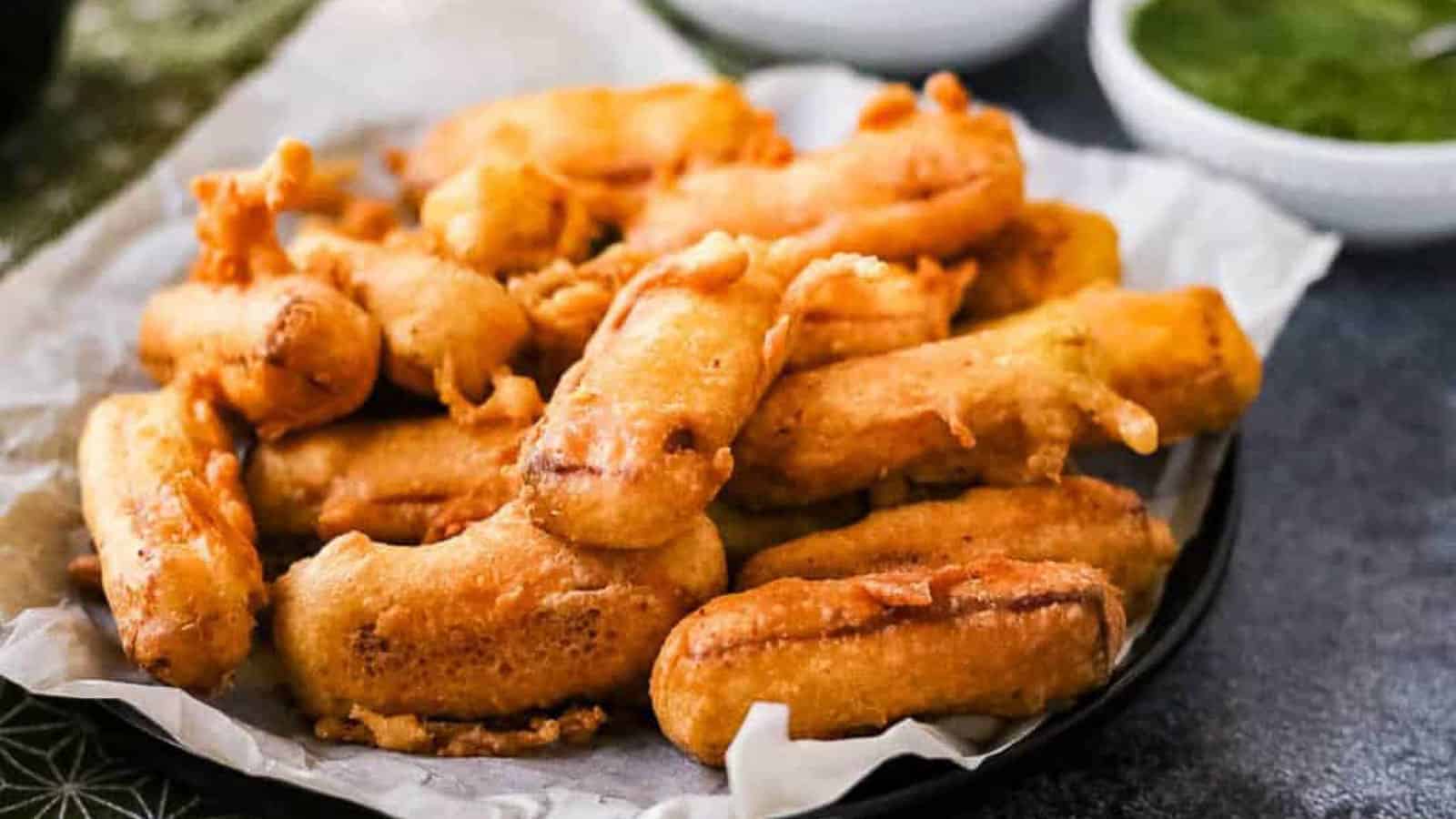 A plate of golden-brown fried snacks on parchment paper. The snacks appear crispy, arranged on a dark circular plate. In the background, there is a small bowl containing green sauce.