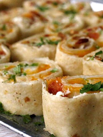 Close-up of a tray filled with rolled tortilla pinwheels. Each pinwheel contains an assortment of ingredients like cheese, bell peppers, and bacon, sprinkled with chopped cilantro. The pinwheels are neatly arranged in rows.