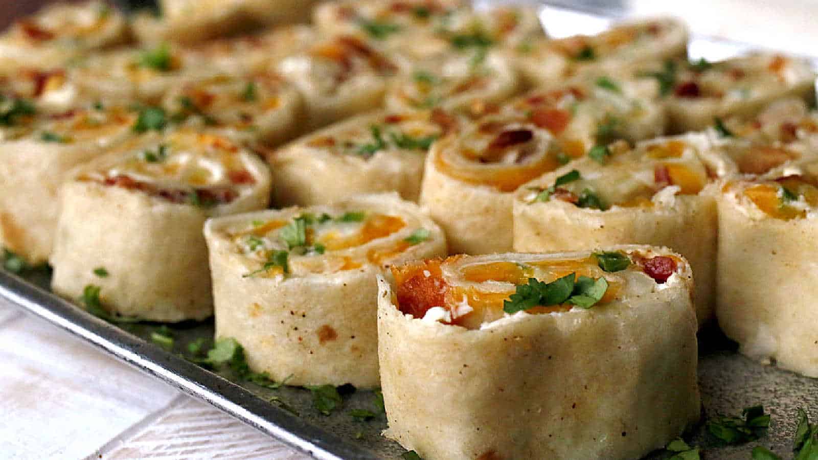 Close-up of a tray filled with rolled tortilla pinwheels. Each pinwheel contains an assortment of ingredients like cheese, bell peppers, and bacon, sprinkled with chopped cilantro. The pinwheels are neatly arranged in rows.