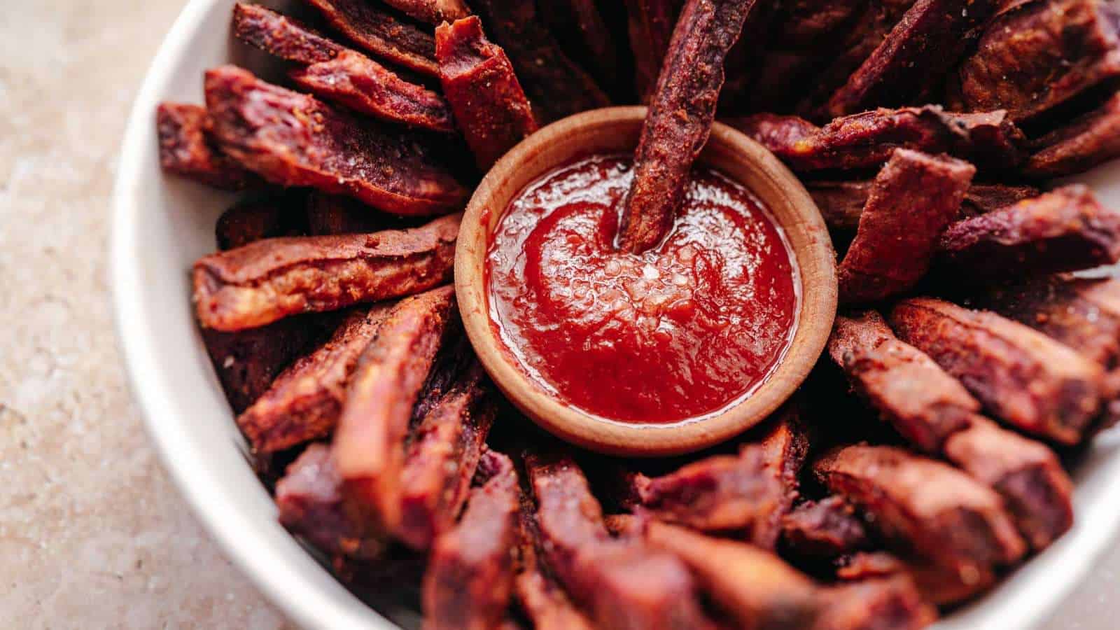 A bowl of crispy sweet potato fries arranged in a circular pattern around a small dip bowl filled with ketchup, placed on a light-colored surface.