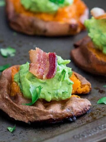 Sweet potato slices topped with mashed avocado, bacon pieces, and chopped herbs are arranged on a baking tray. The surface of the tray is slightly worn, and the toppings are vibrant in color.