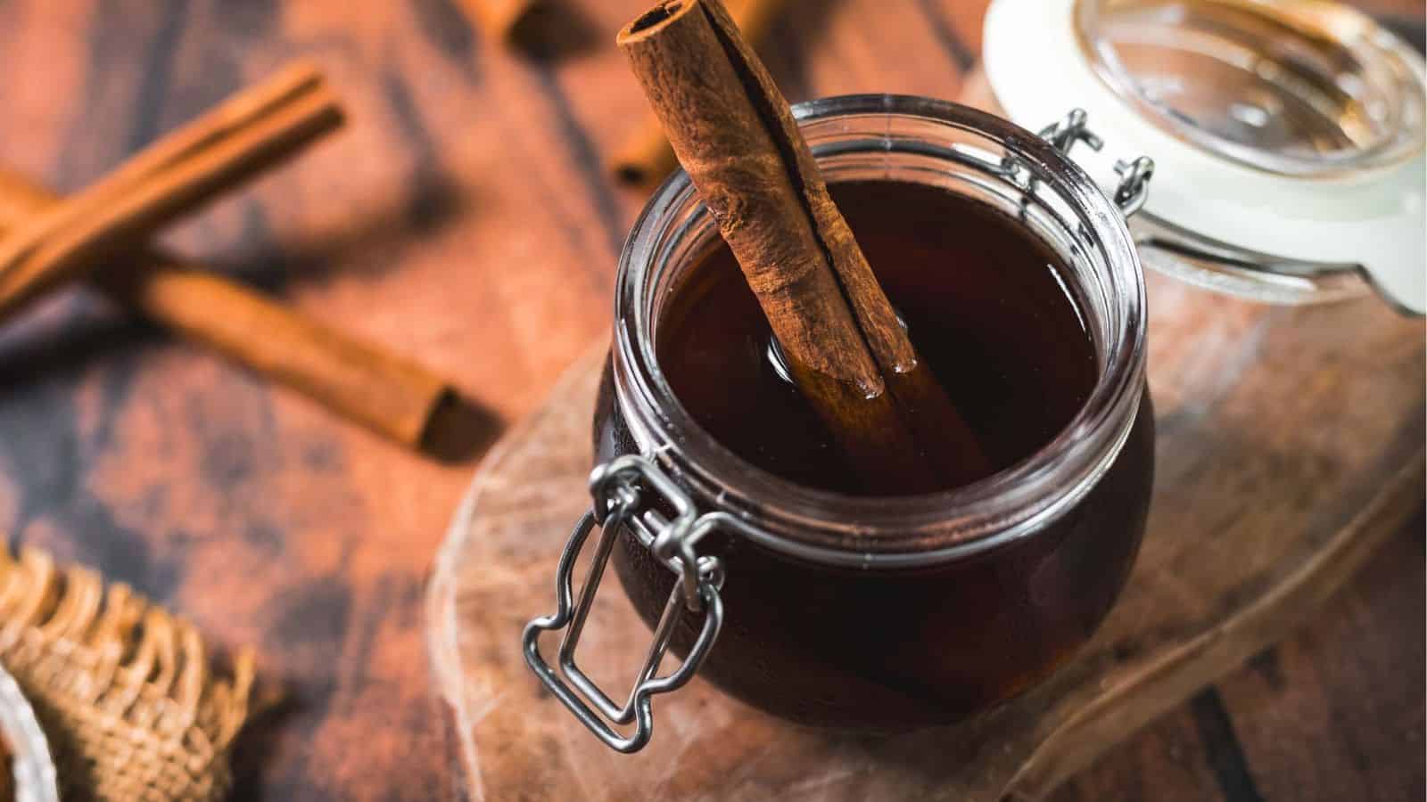 A glass jar filled with dark liquid, likely cinnamon-infused, sits on a wooden surface. A cinnamon stick is placed inside the jar. The jar has a metal clasp and white lid. Nearby, more cinnamon sticks are scattered on the table.
