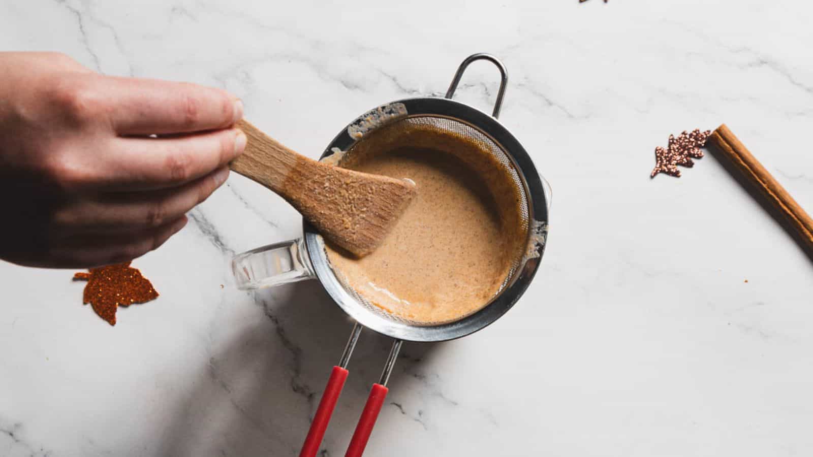 A hand uses a wooden spatula to stir a creamy mixture in a glass measuring cup placed over a metal strainer. The setup is on a white marble surface, with small decorative leaves and a cinnamon stick nearby.