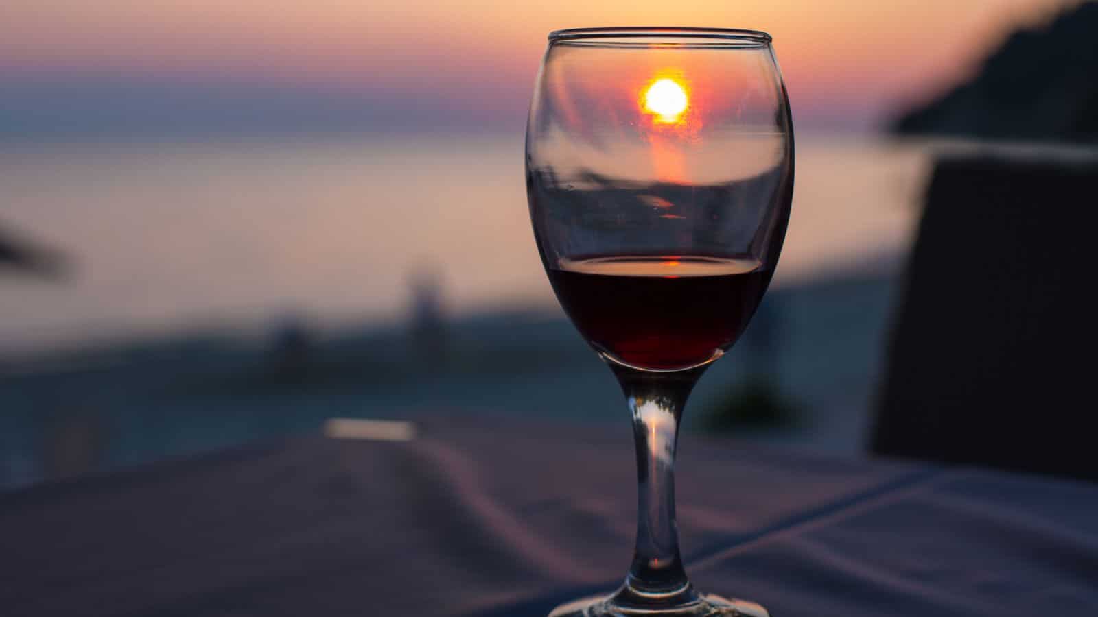 A glass of red wine sits on a table overlooking a beach at sunset. The sun is positioned through the wine glass, casting a warm glow. The ocean and sky create a calm backdrop with hues of pink and orange.