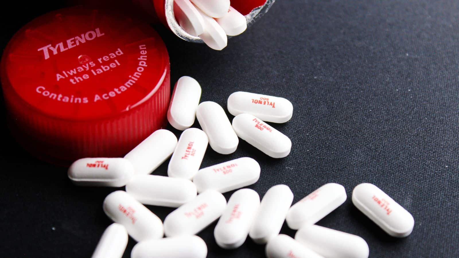 A red Tylenol bottle cap lies open on a black surface, with white caplets labeled "Tylenol" scatter around it. The cap reads "Always read the label" and "Contains Acetaminophen.
