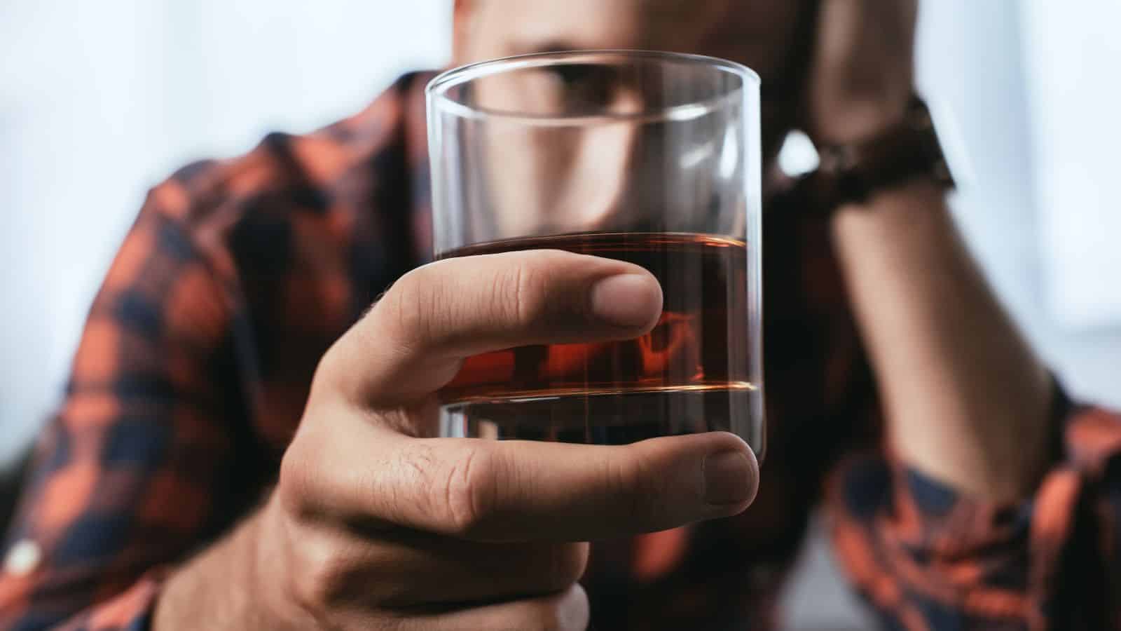 A person in a plaid shirt holds a glass of dark liquid, possibly whiskey, close to their face. The focus is on the hand and glass, while the person's face and surroundings are blurred.