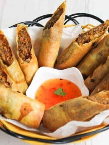 A black wire basket holds several fried spring rolls with a meat filling, each cut in half diagonally. In the center is a small white dish containing orange dipping sauce. A red and white checkered cloth is partially visible on the side.