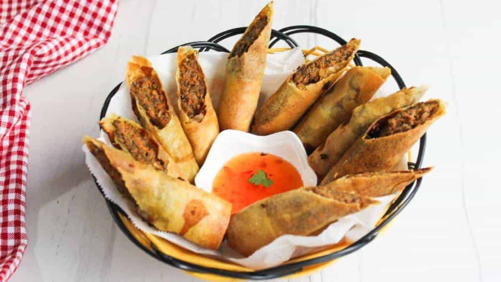 A black wire basket holds several fried spring rolls with a meat filling, each cut in half diagonally. In the center is a small white dish containing orange dipping sauce. A red and white checkered cloth is partially visible on the side.