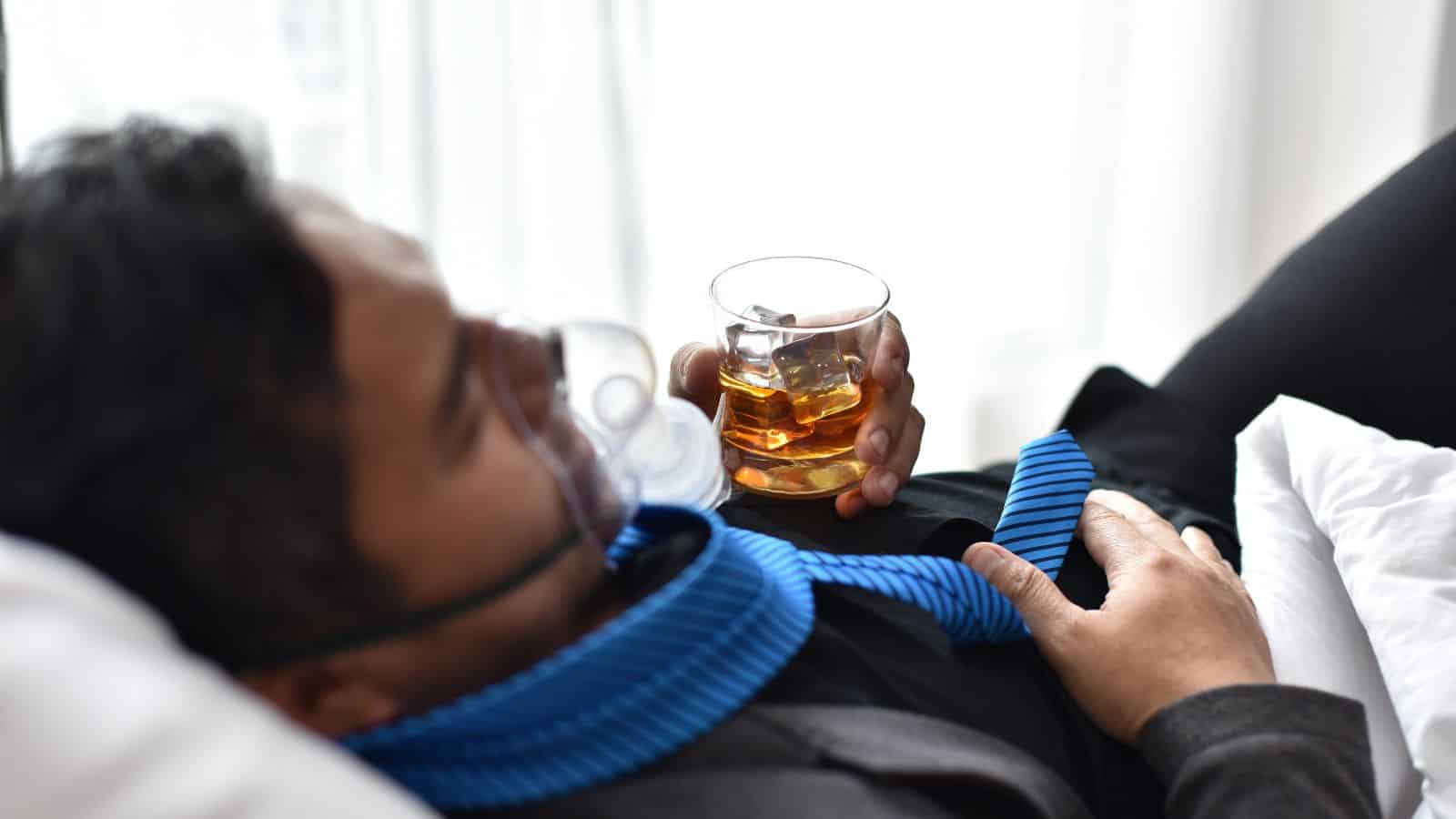 A person is reclining on a bed, wearing an oxygen mask, holding a glass of amber liquid with ice cubes. They have a blue tie loosely draped around their neck and are dressed in black attire. Soft light filters through a window behind them.