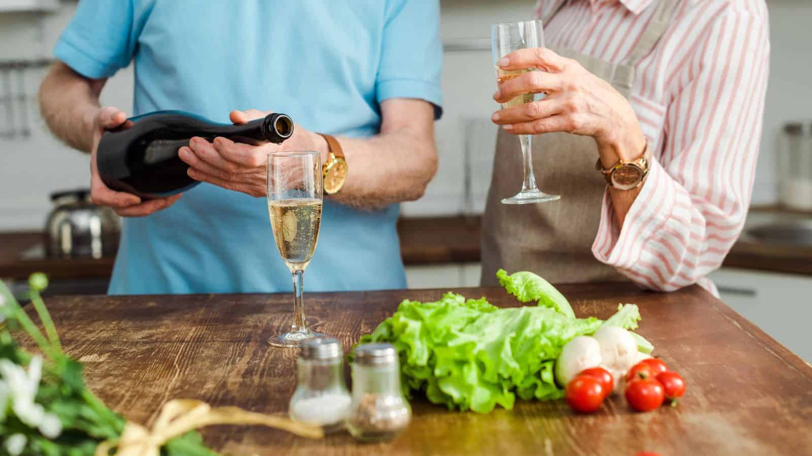 Two people stand at a kitchen counter. One holds a glass of champagne while the other pours more into a glass. Fresh vegetables, including lettuce and tomatoes, are on the counter. They both wear watches and casual clothing.