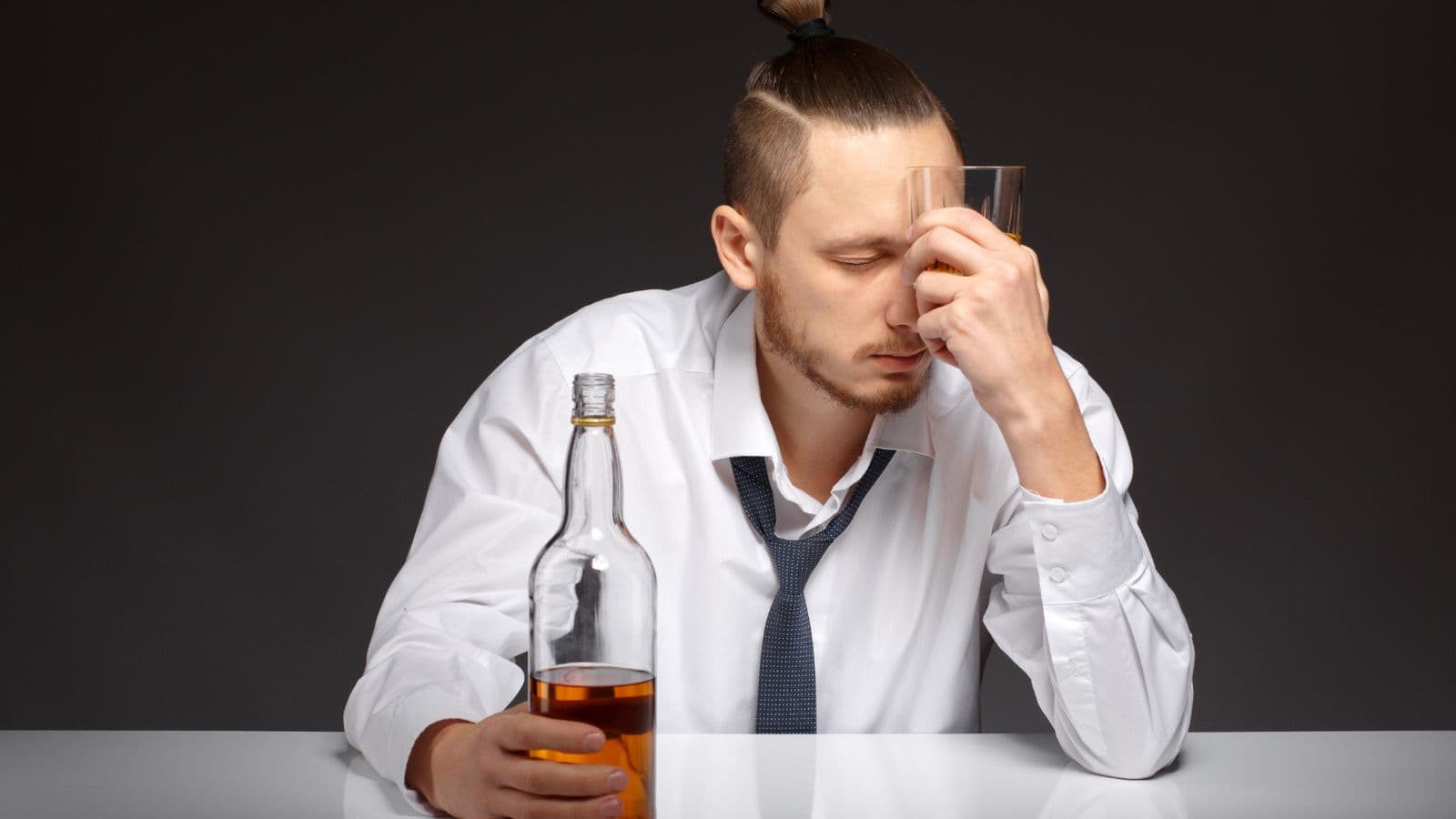 A person in a white shirt and tie, holding a glass to their forehead, sits at a table with a bottle of amber liquid in the other hand. The background is dark, and their expression appears contemplative or stressed.