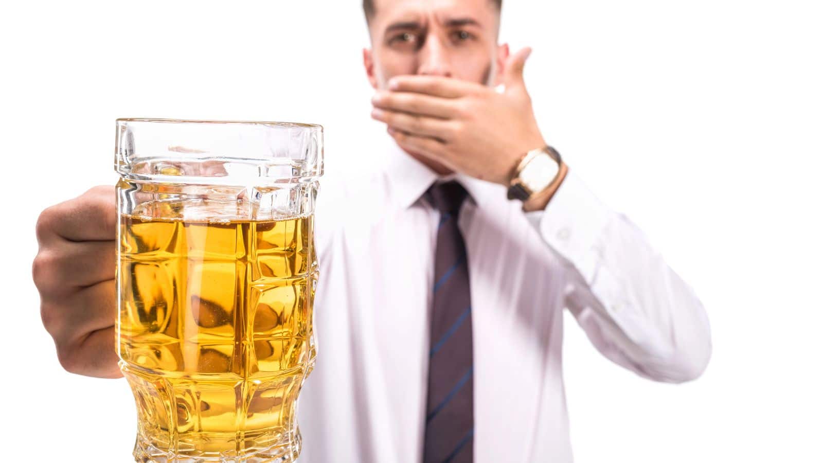 A person in a shirt and tie covers their mouth with one hand while holding a glass mug filled with a light-colored beverage in the other hand, against a plain white background.