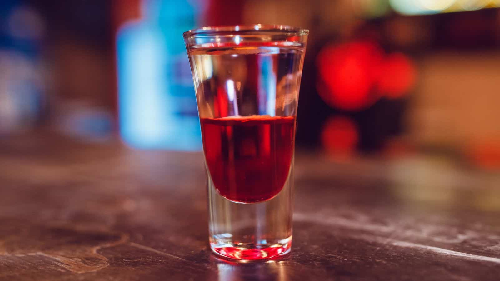 A layered shot cocktail sits on a bar counter. The glass contains a red liquid on top of a clear liquid, creating a visible separation. The background is blurred, featuring warm, ambient lighting.
