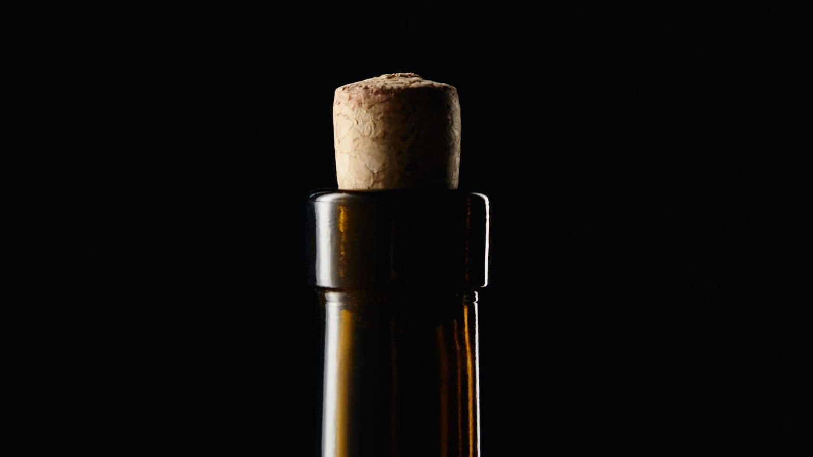 A close-up image of the neck of a dark brown glass bottle with a cork stopper. The background is black, highlighting the bottle and cork.