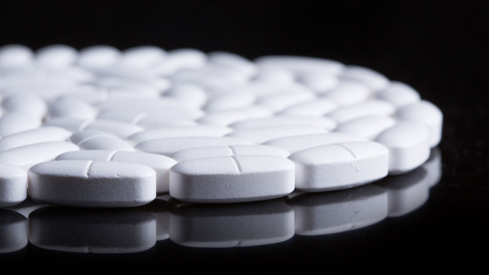 A close-up image shows numerous white oval tablets, each with a cross-shaped score line, arranged in a circular pattern on a reflective black surface. The focus is on the tablets in the foreground.