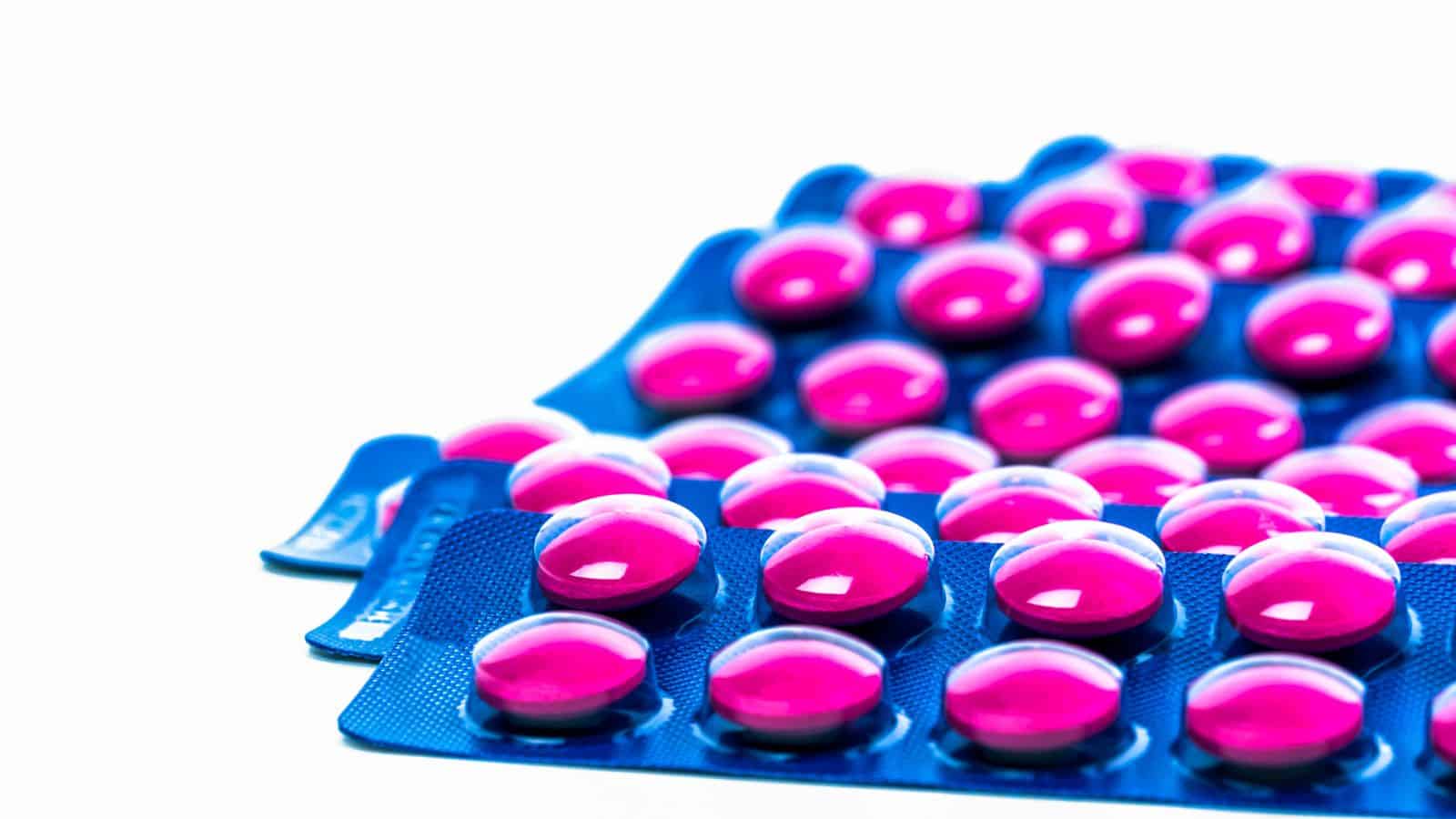 Blister packs filled with round, pink pills are arranged in overlapping rows against a white background. The packaging is blue, and the pills are glossy and evenly spaced.