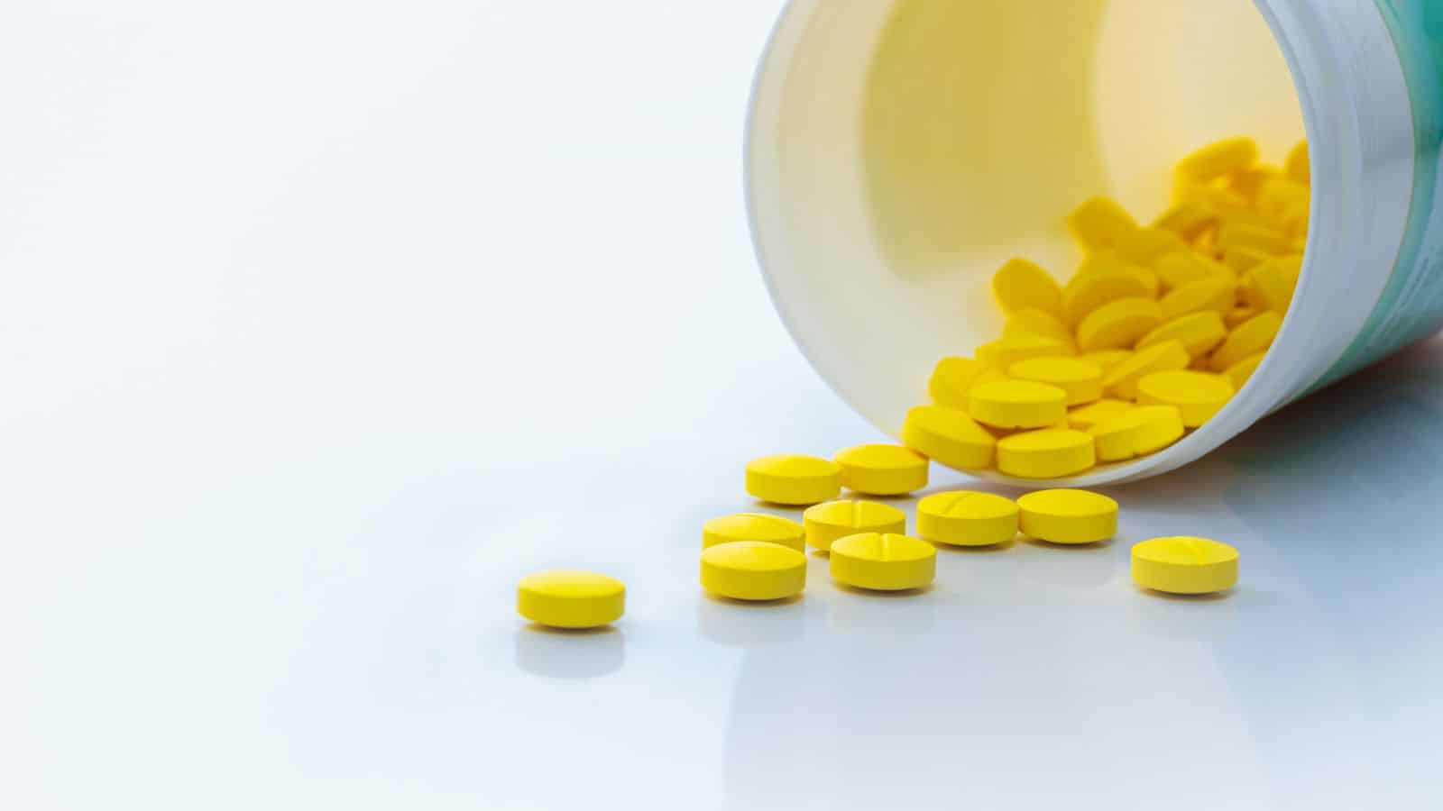 A white container lies on its side, spilling out numerous round yellow tablets onto a smooth white surface. The tablets are scattered in front of the open container.
