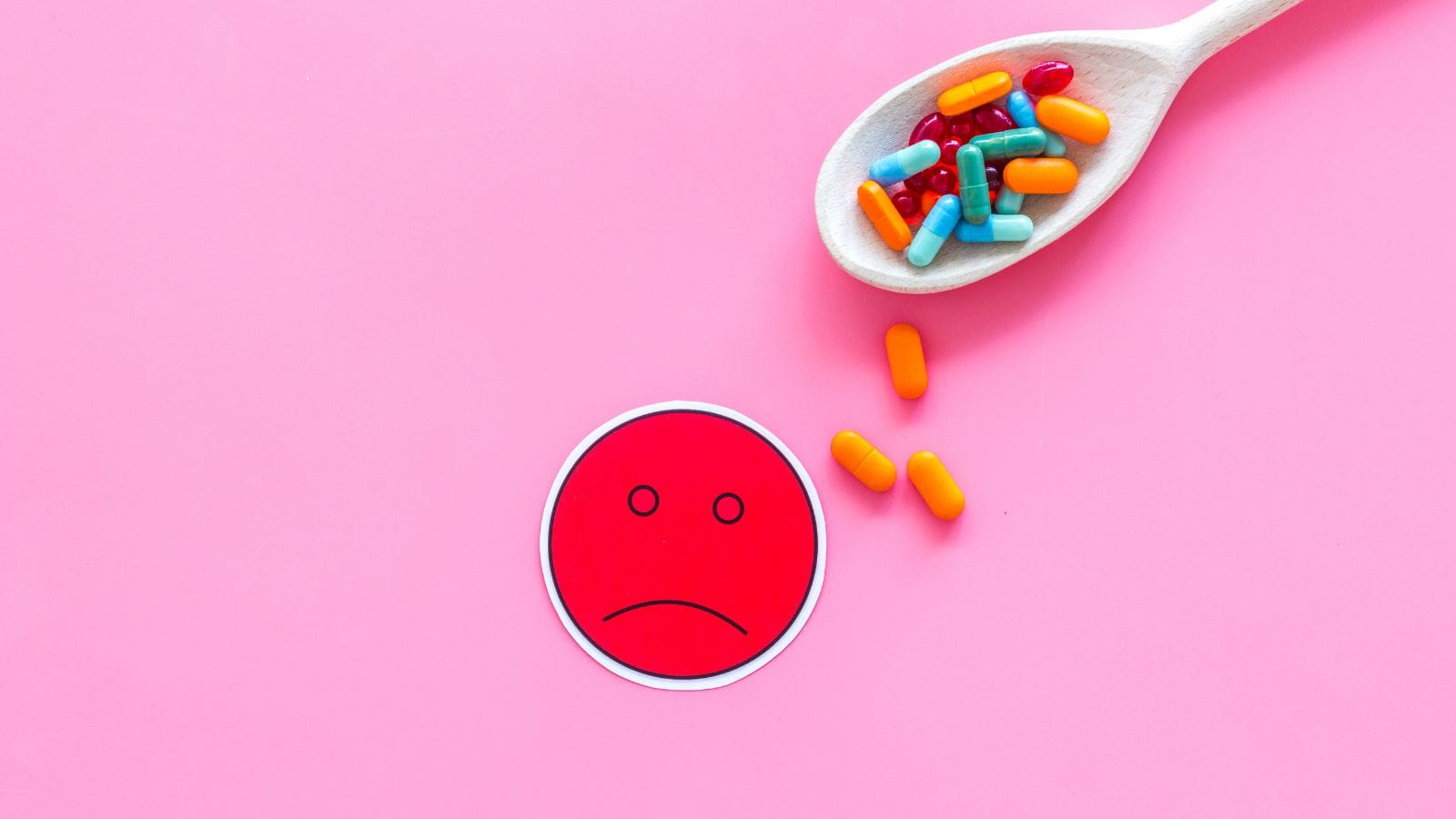 A wooden spoon holds assorted colorful capsules and pills on a pink background. A red paper circle with an unhappy face is placed nearby.