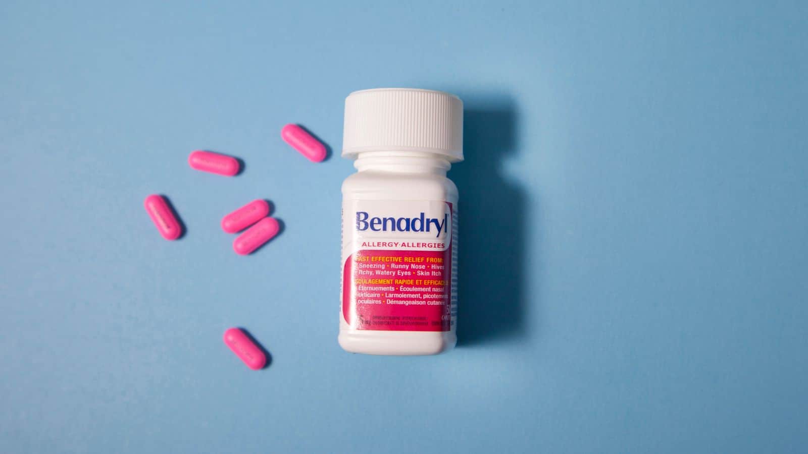 A bottle of Benadryl Allergy tablets is positioned on a blue background, with six pink tablets scattered to the left of the bottle. The bottle has a white cap and a red label with text indicating the medication's use.