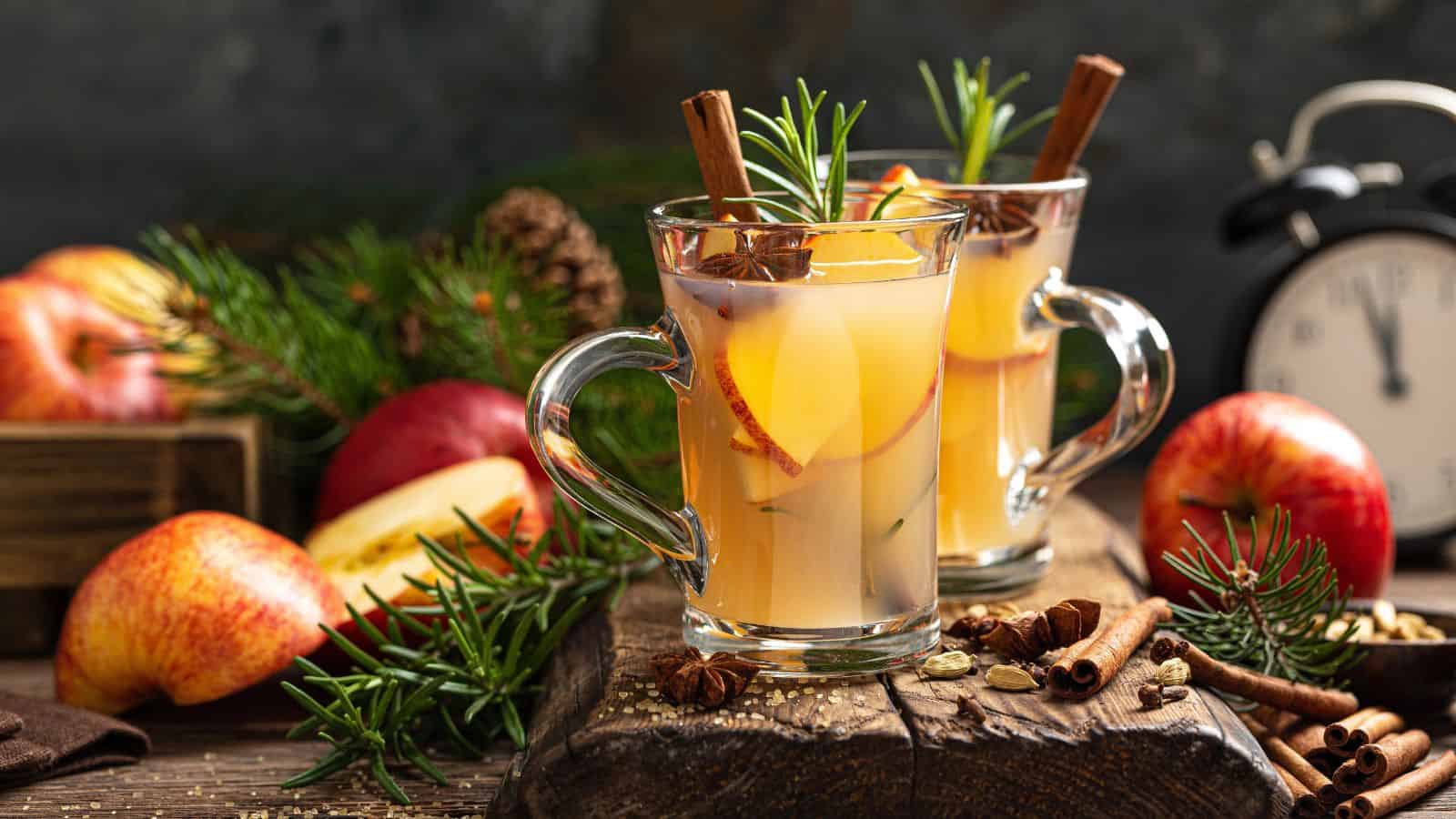 Two glass mugs filled with a warm beverage, garnished with apple slices, cinnamon sticks, and rosemary sprigs. Surrounding the mugs are apples, pine cones, an alarm clock, and cinnamon sticks, all set on a wooden surface.