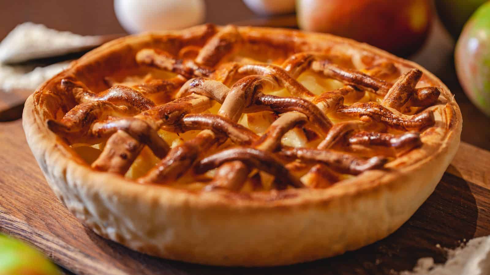A freshly baked apple pie with a golden brown lattice crust sits on a wooden board. In the background, there are blurred apples and ingredients on a table.