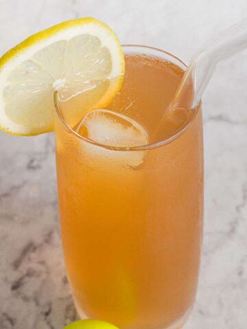 A tall glass of iced peach tea on a marble surface, garnished with a lemon slice and a clear straw.