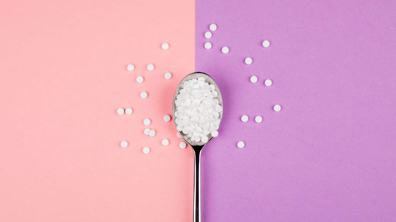 A spoon filled with small white pills or tablets is placed on a flat surface, divided into two halves: pink on the left and purple on the right. Some pills are scattered around the spoon on both sides of the surface.