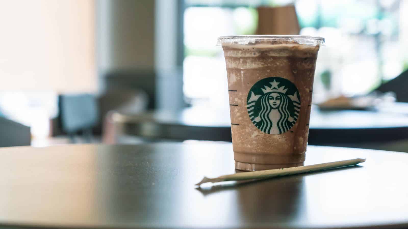 A Starbucks plastic cup filled with a blended iced beverage sits on a table. A green straw lies next to the cup. The background shows blurred indoor seating with soft lighting.