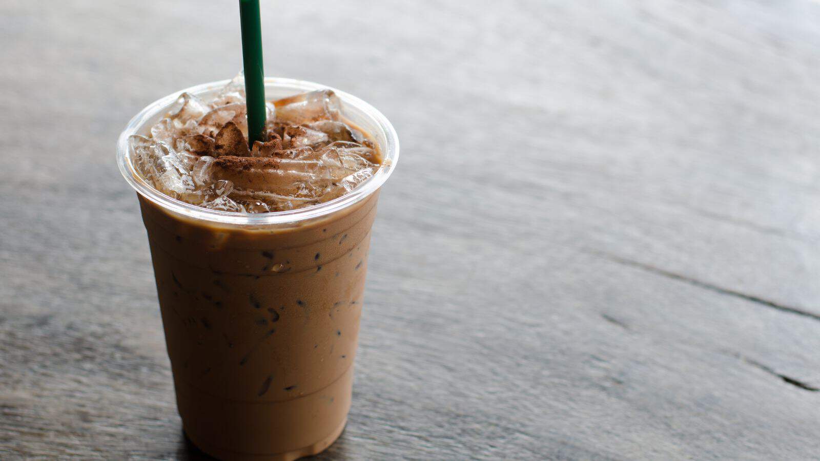 A plastic cup filled with iced coffee sits on a wooden surface. The drink has a green straw and is topped with ice cubes.