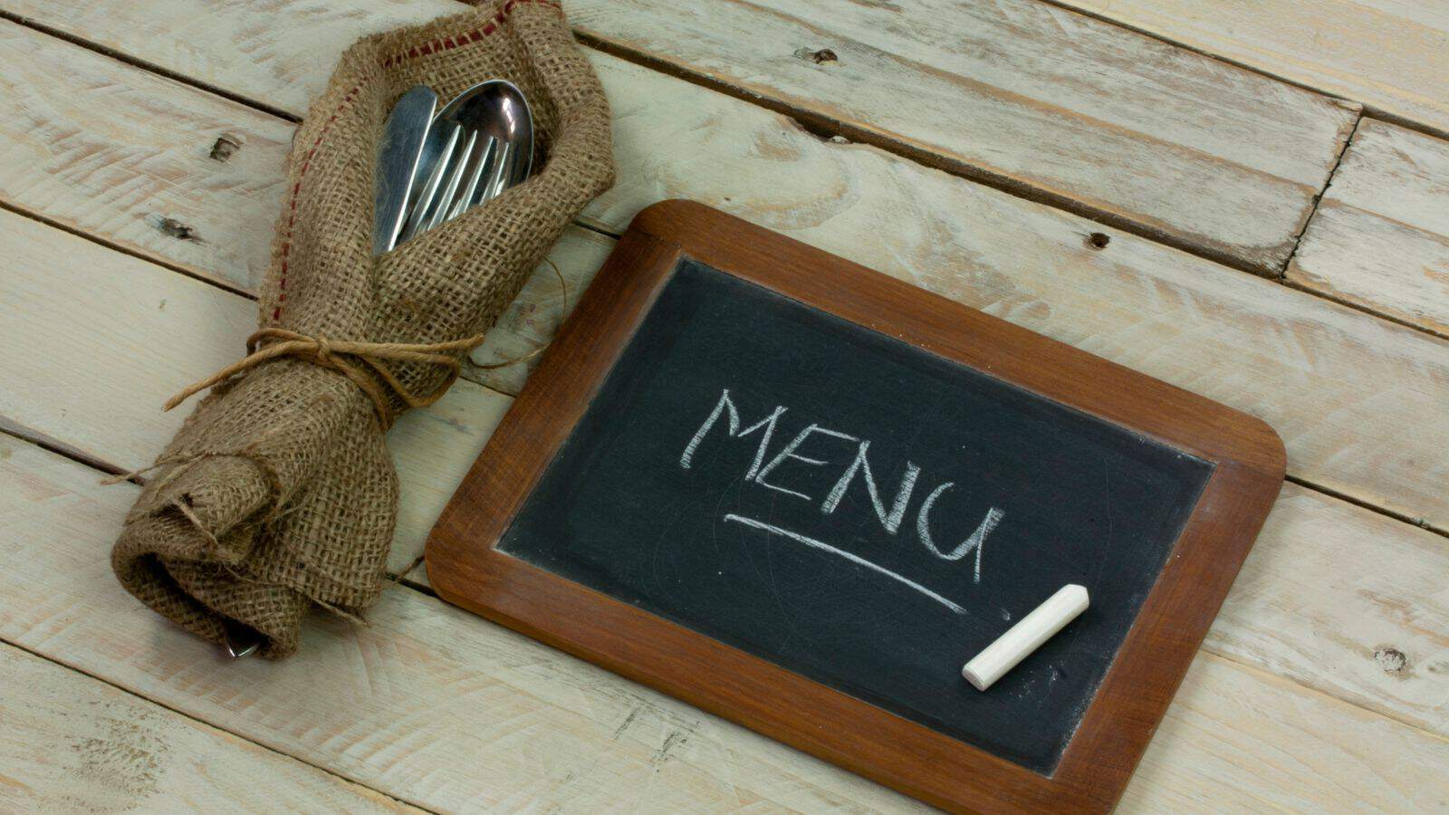 A small chalkboard with "MENU" written in white chalk rests on a wooden table. Next to it, cutlery wrapped in burlap tied with twine is placed on the same surface.