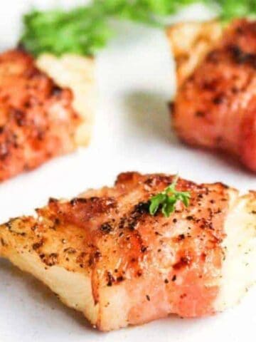 Three pieces of cooked bacon-wrapped chicken are garnished with parsley on a white surface. The chicken appears seasoned with black pepper.