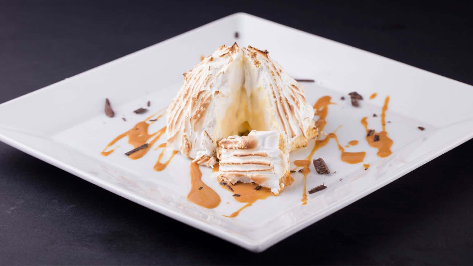 A baked Alaska dessert is served on a white square plate against a dark background. The dessert features a browned meringue exterior, a drizzle of caramel sauce, and scattered chocolate shavings. A section is cut to reveal the inside.