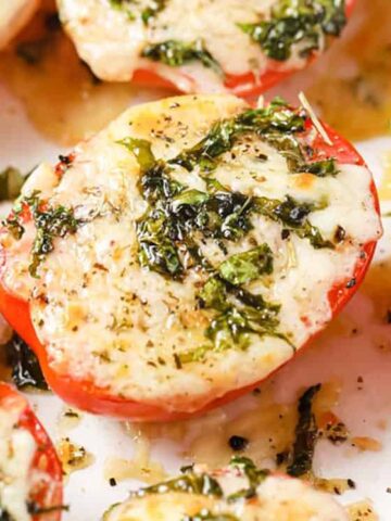 Sliced tomatoes topped with melted cheese, herbs, and seasoning are laid out on a baking sheet. The cheese is lightly browned, and the herbs are green, adding texture and color to the dish.