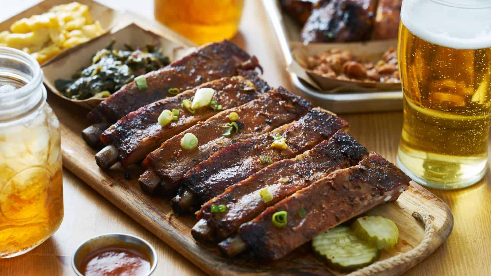 A wooden platter with grilled ribs garnished with green onions, accompanied by pickles. Sides of greens and scrambled eggs are in the background. Two glasses of beer are nearby, and a jar with a cold beverage sits to the left.