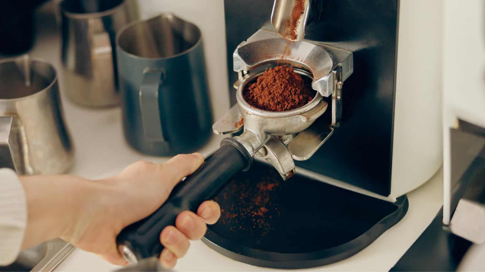 A person holds a portafilter filled with ground coffee under a coffee grinder. Several metal pitchers are in the background, placed on a white surface. The image focuses on the preparation of coffee grounds for espresso.