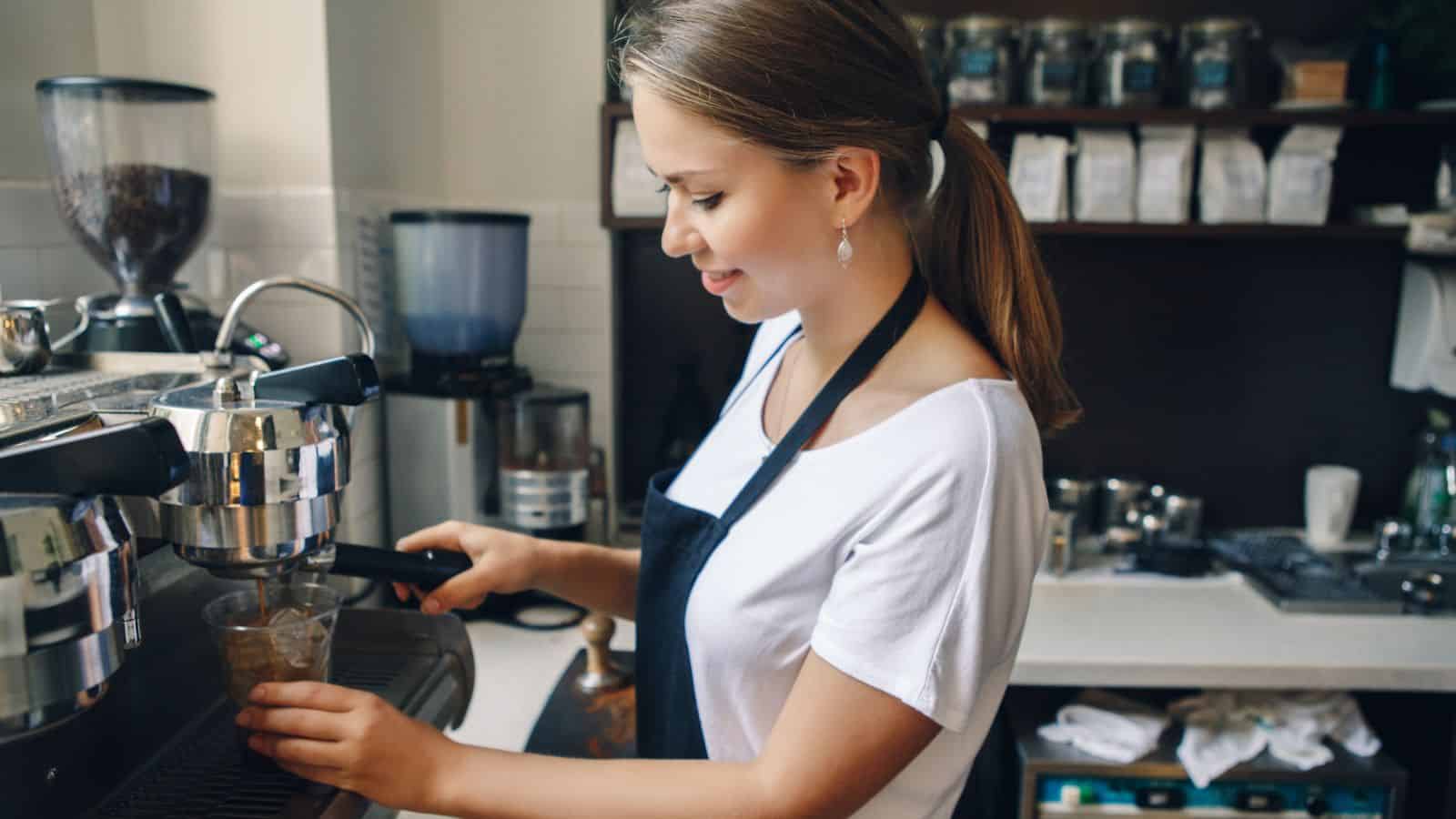 A person in a white shirt and black apron is preparing a coffee using an espresso machine. They are in a coffee shop, with shelves in the background holding coffee bags and jars.
