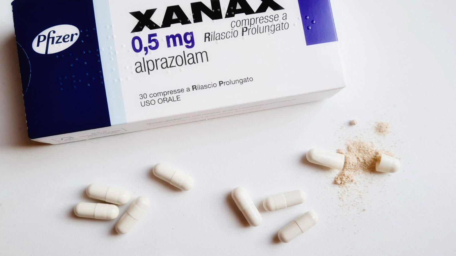 A box of Xanax (alprazolam) 0.5 mg tablets is shown with Pfizer branding. Several white capsules are scattered in front of the box, along with some powder on a white surface.