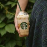 A person holding a Starbucks cup filled with a caramel-colored frappuccino topped with whipped cream and drizzled syrup, against a backdrop of green leaves. The person is wearing a plaid skirt.