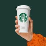 Hand holding a white Starbucks coffee cup with the green logo on the front. The person is wearing an orange knitted sweater. The background is solid dark green.