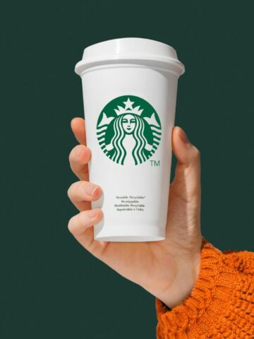 Hand holding a white Starbucks coffee cup with the green logo on the front. The person is wearing an orange knitted sweater. The background is solid dark green.