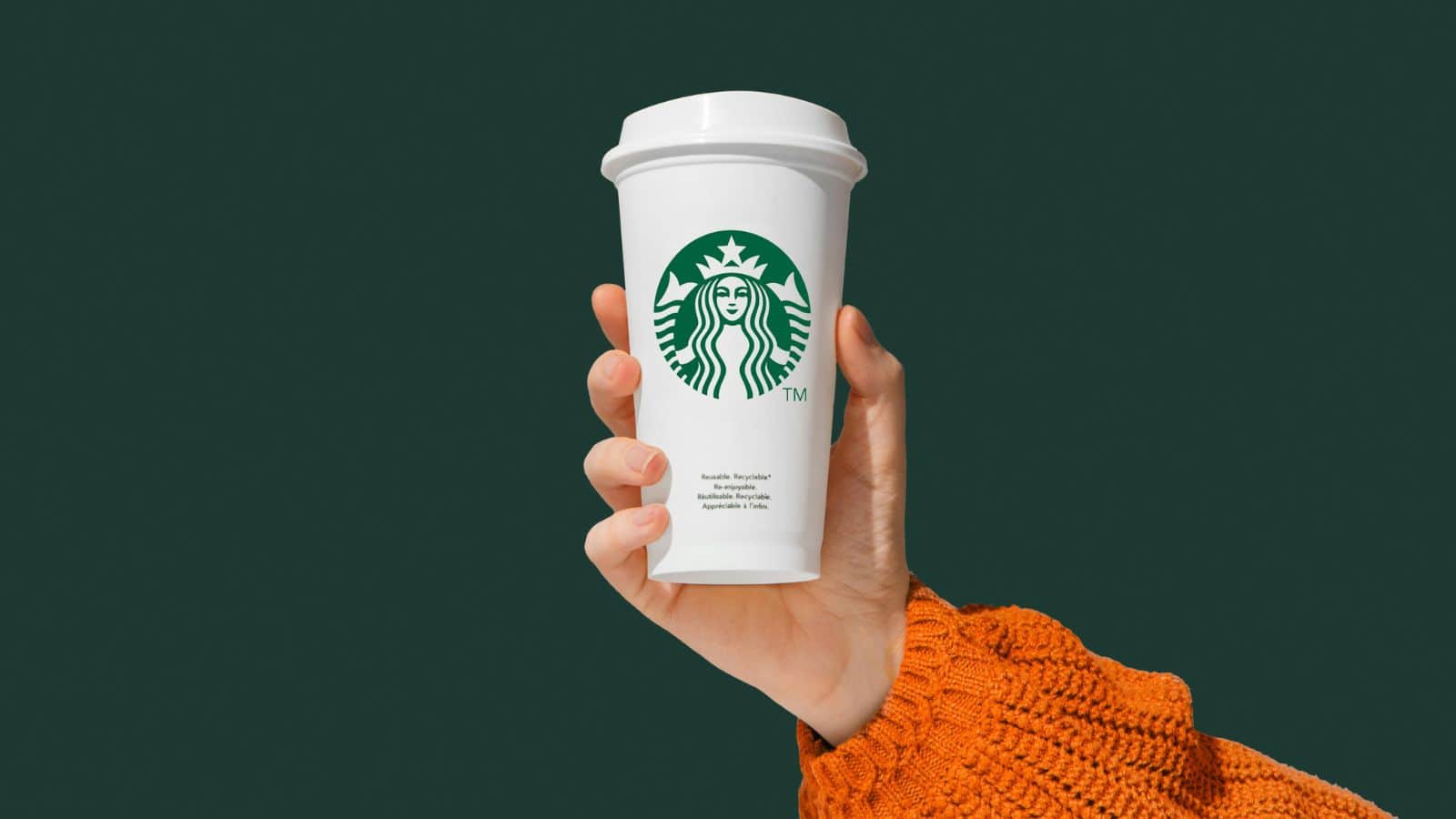 Hand holding a white Starbucks coffee cup with the green logo on the front. The person is wearing an orange knitted sweater. The background is solid dark green.