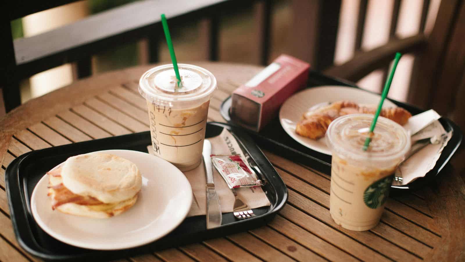 Two trays on a wooden table, each with a cold drink in a plastic cup with a green straw. One tray has a sandwich and a wrapped snack. The other tray holds a boxed pastry and a croissant on a plate. Napkins and cutlery are visible.