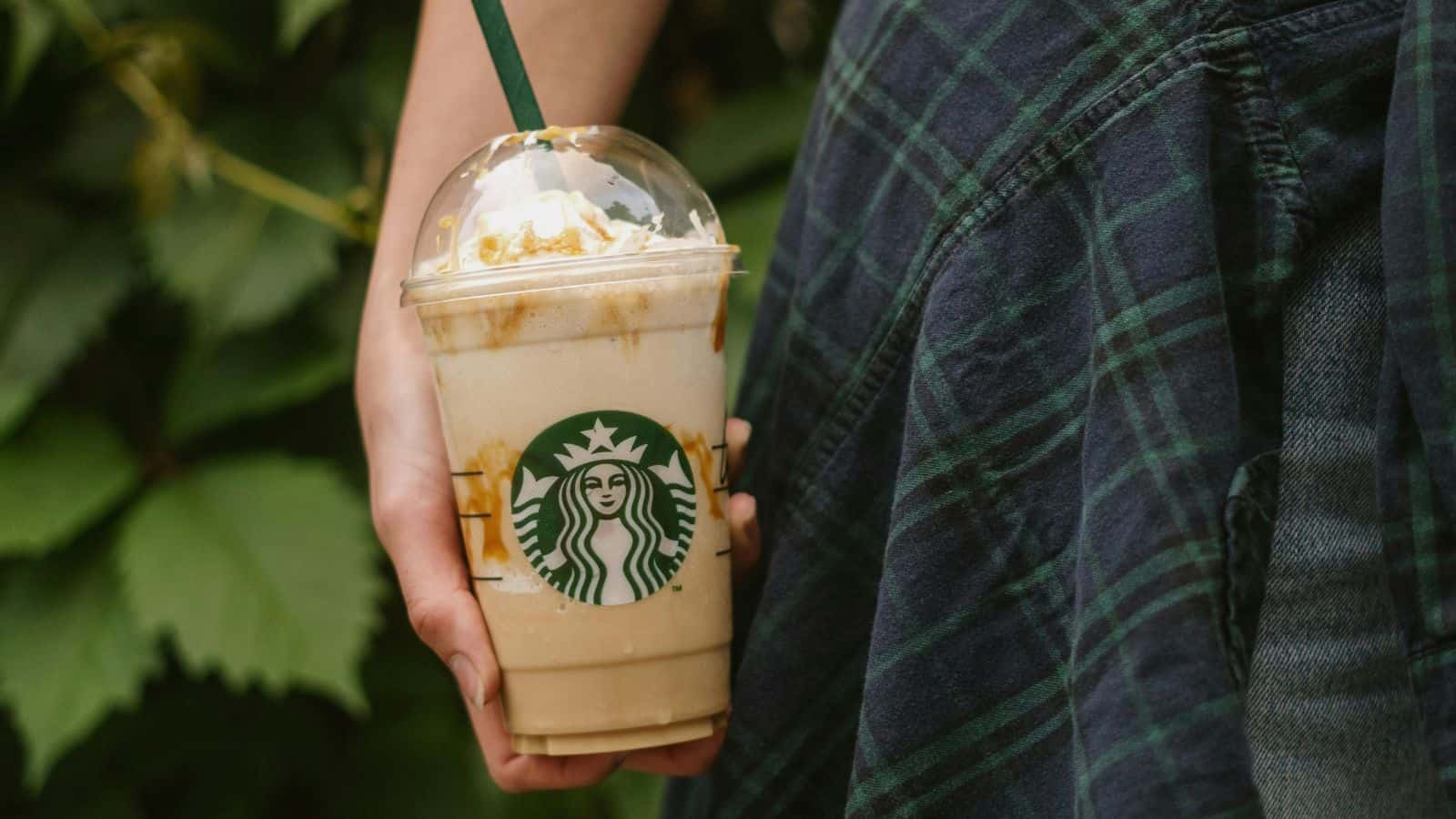 A person is holding a Starbucks Frappuccino with caramel drizzle and whipped cream. The cup is clear with a dome lid and green straw. The person is wearing a plaid garment, and green leaves are visible in the background.
