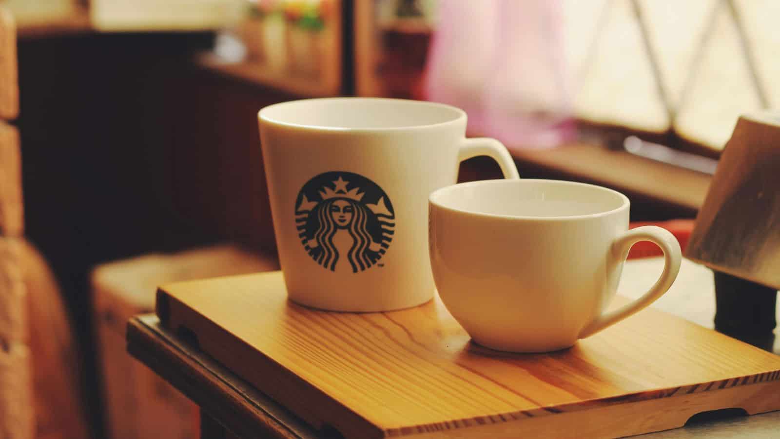 Two white ceramic coffee mugs are placed on a wooden tray. The larger mug displays the Starbucks logo. Sunlight filters through a window in the background, creating a warm and cozy atmosphere.