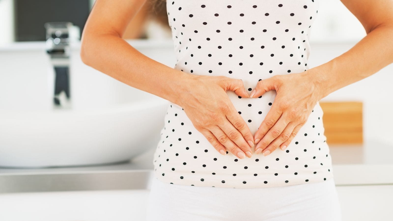 A person wearing a white tank top with black polka dots shapes their hands into a heart over their stomach. A bathroom sink and counter are visible in the background.