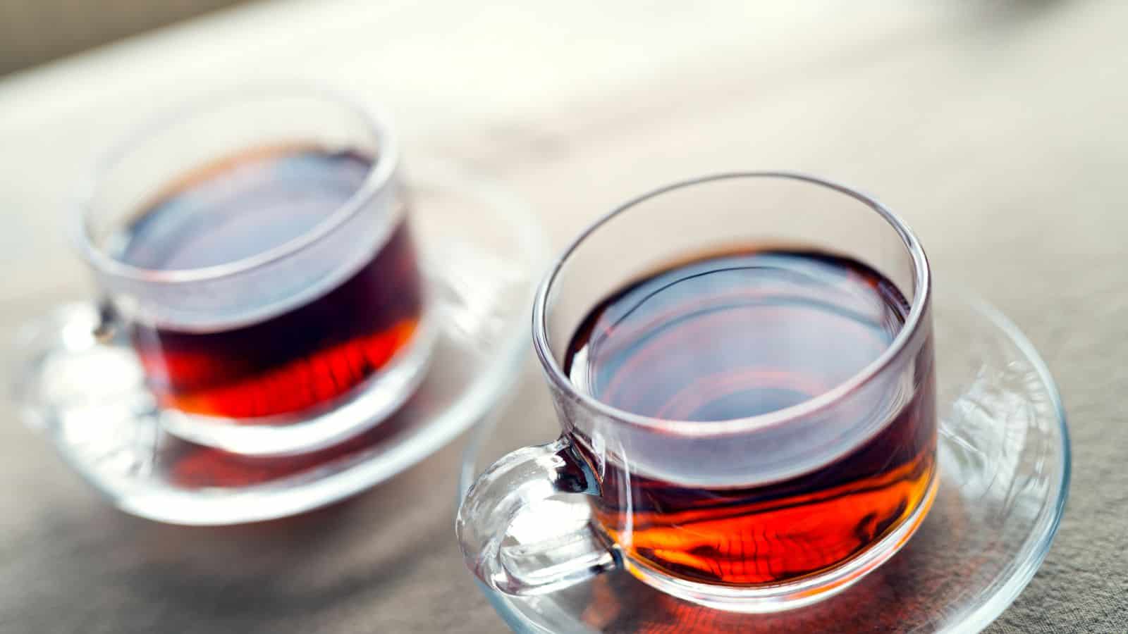 Two clear glass cups filled with dark tea are placed on matching saucers on a light surface. The tea has a reddish-brown hue, and the glass cups show reflections on their sides. The background is softly blurred.
