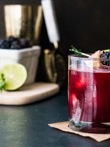 A glass filled with a dark red cocktail garnished with blackberries and a sprig of fresh mint, sitting on a wooden coaster. In the background, a lime, mint, and a container of blackberries are placed on a cutting board.