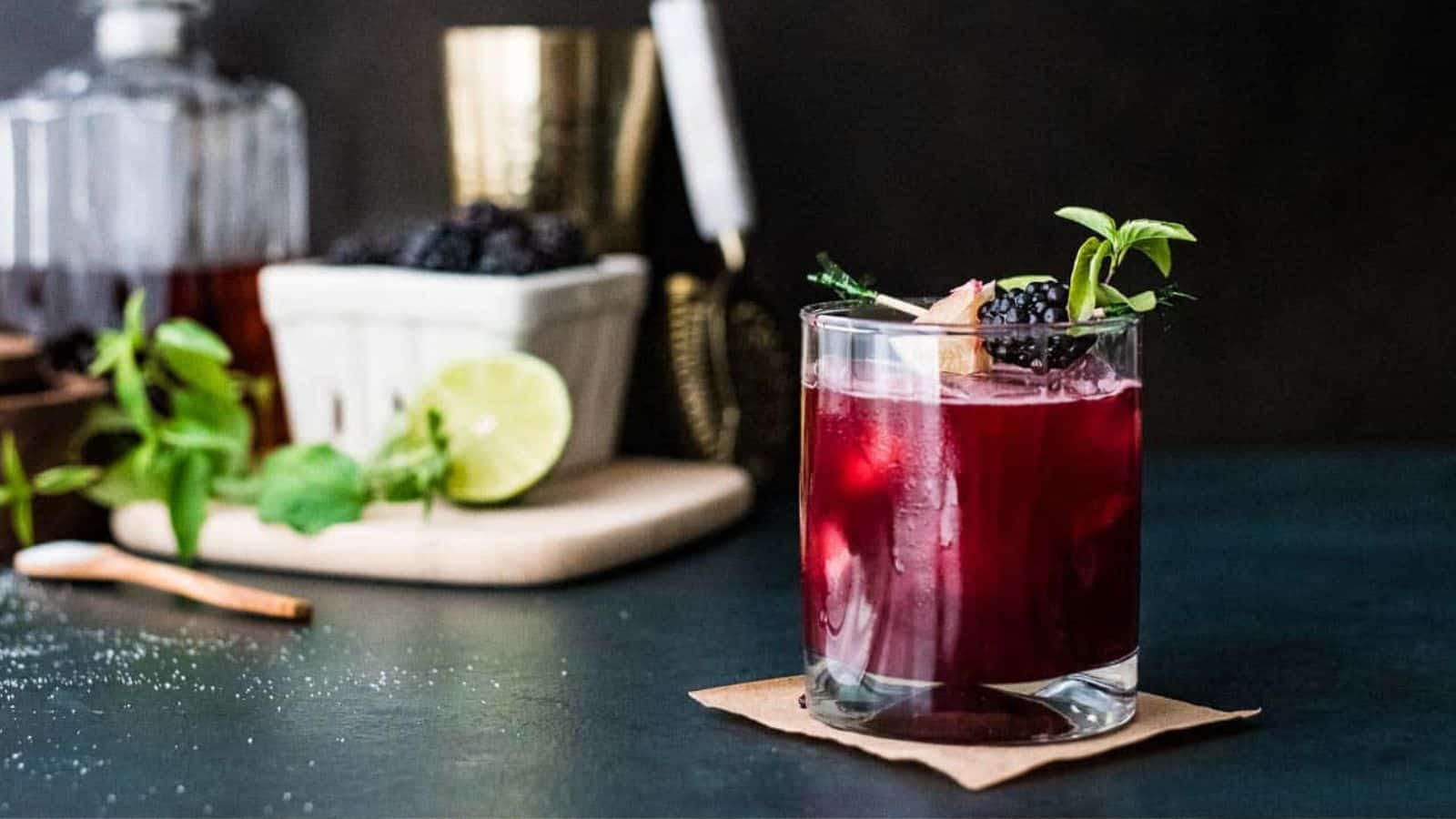 A glass filled with a dark red cocktail garnished with blackberries and a sprig of fresh mint, sitting on a wooden coaster. In the background, a lime, mint, and a container of blackberries are placed on a cutting board.