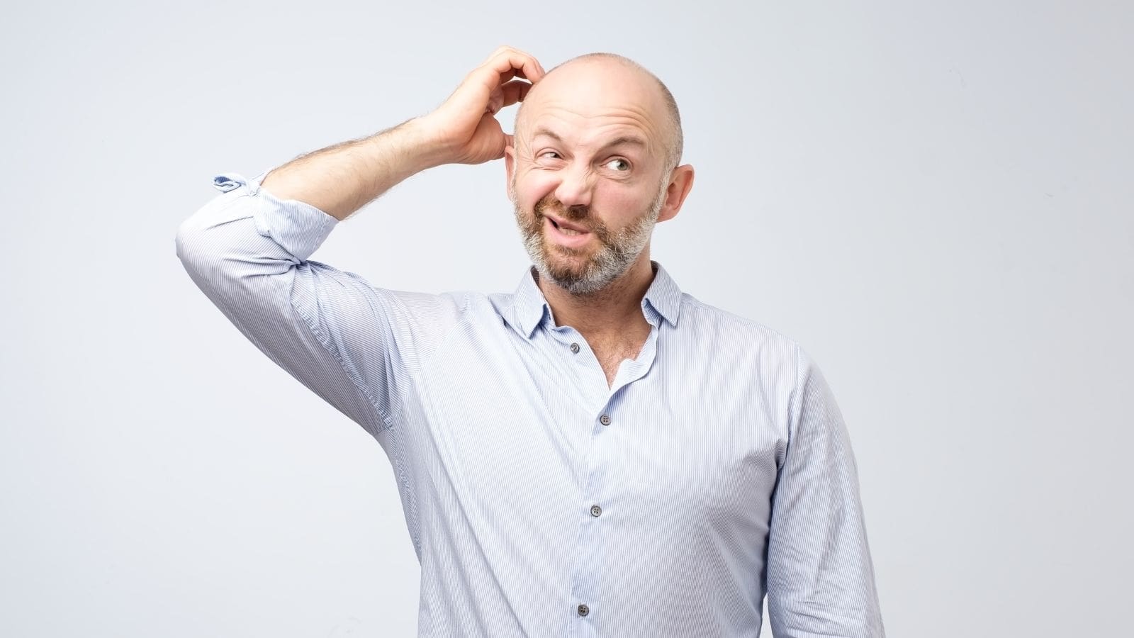 A bald man with a beard is scratching his head and making a confused expression. He is wearing a light blue button-up shirt and is set against a plain white background.